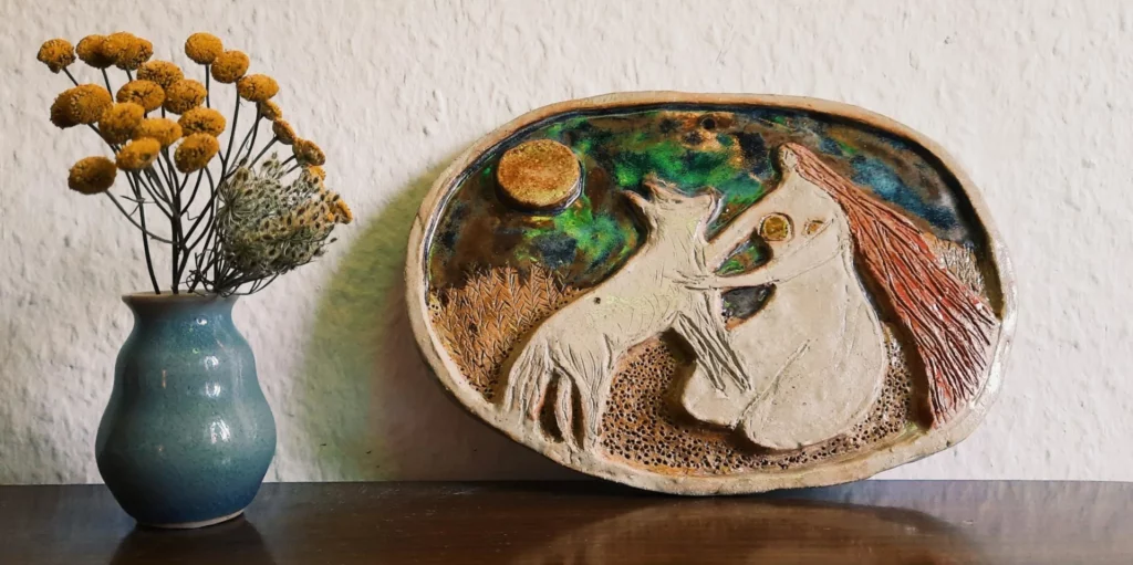 wolf woman - a ceramic relief showcasing a kneeling woman with long hair in a natural setting, holding a wolf on her lap, its head touching her face. Above them is a colourful, whimsical sky with a full moon, symbolising female wilderness and a deep connection to natural cycles and power.